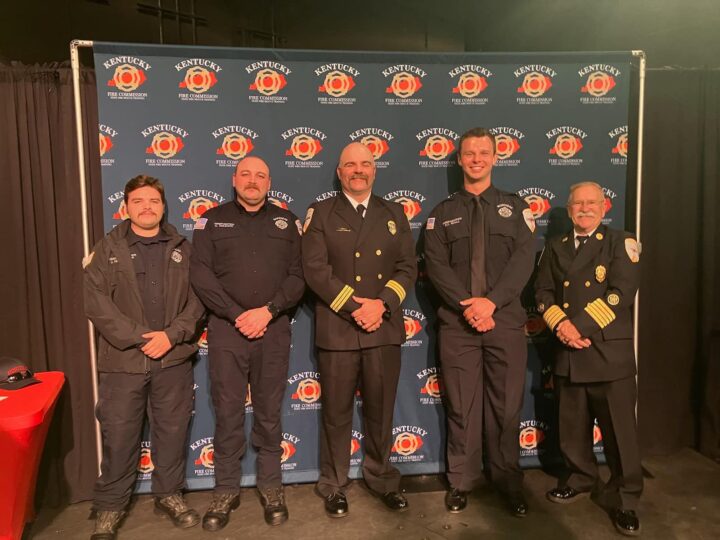 “Five Morehead firefighters and fire department officials in uniform pose in front of a Kentucky Fire Commission banner at an academy graduation.”