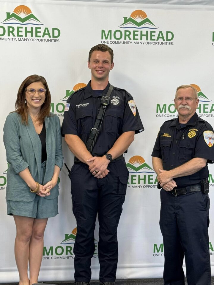 “Firefighter CJ Wall stands between the mayor and the fire chief in front of a Morehead logo backdrop, all smiling at the camera.”