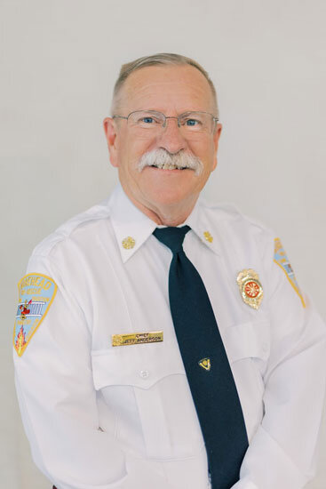“Older man with short light hair, glasses, and a white mustache, wearing a white fire-department uniform and dark tie, smiling in front of a light background.”