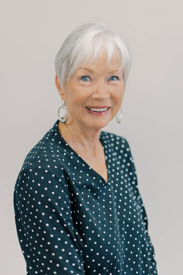 “Older woman with short white hair, wearing a dark blouse with white polka dots and hoop earrings, smiling in front of a light background.”