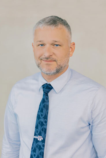 “Man with short gray hair and a trimmed beard, wearing a light blue dress shirt and a dark patterned tie, smiling against a light background.”