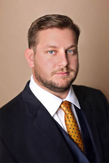 “Man with short light-brown hair and trimmed beard, dressed in a dark suit with a white shirt and yellow patterned tie, facing the camera with a neutral expression.”