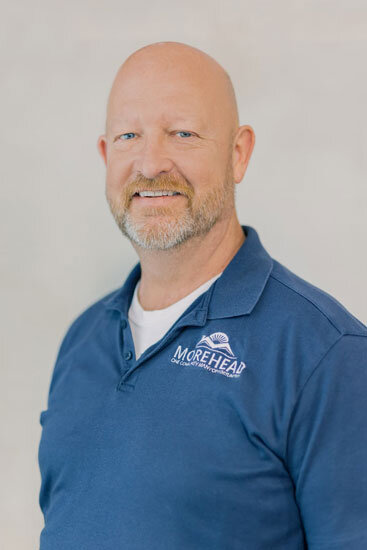 “Bald man with a trimmed beard, wearing a blue collared shirt with a city logo, smiling slightly against a light background.”