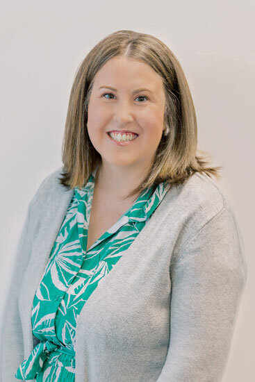 “Woman with shoulder-length light brown hair, wearing a green patterned blouse and light gray cardigan, smiling in front of a white background.”
