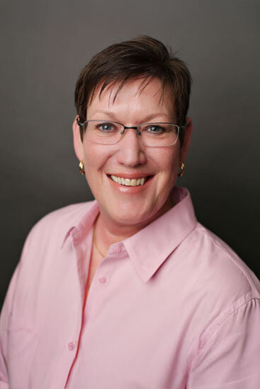 “Woman with short brown hair and glasses, wearing a light pink collared shirt, smiling in front of a dark gray background.”