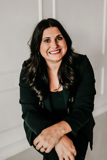 “Woman with long dark wavy hair, wearing a black blazer, seated and smiling warmly in front of a light-colored wall.”