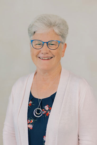 “Older woman with short gray hair and blue glasses, wearing a light pink cardigan over a dark floral top, smiling in front of a light background.”