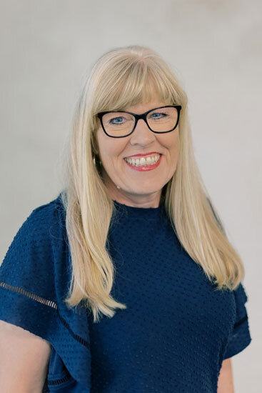 “Woman with long blonde hair and black glasses, wearing a dark blue textured top, smiling against a light background.”