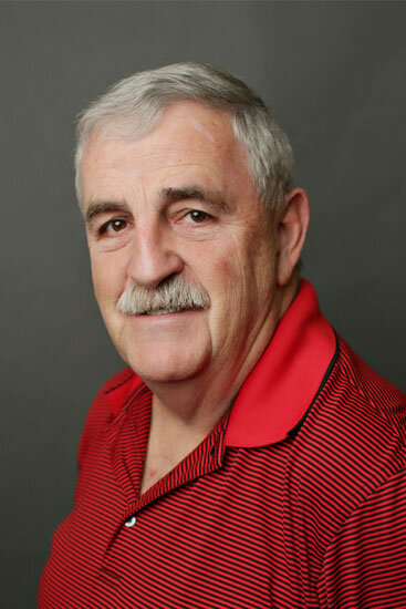 “Older man with short gray hair and a gray mustache, wearing a red collared shirt with thin black stripes, smiling slightly against a plain gray background.”